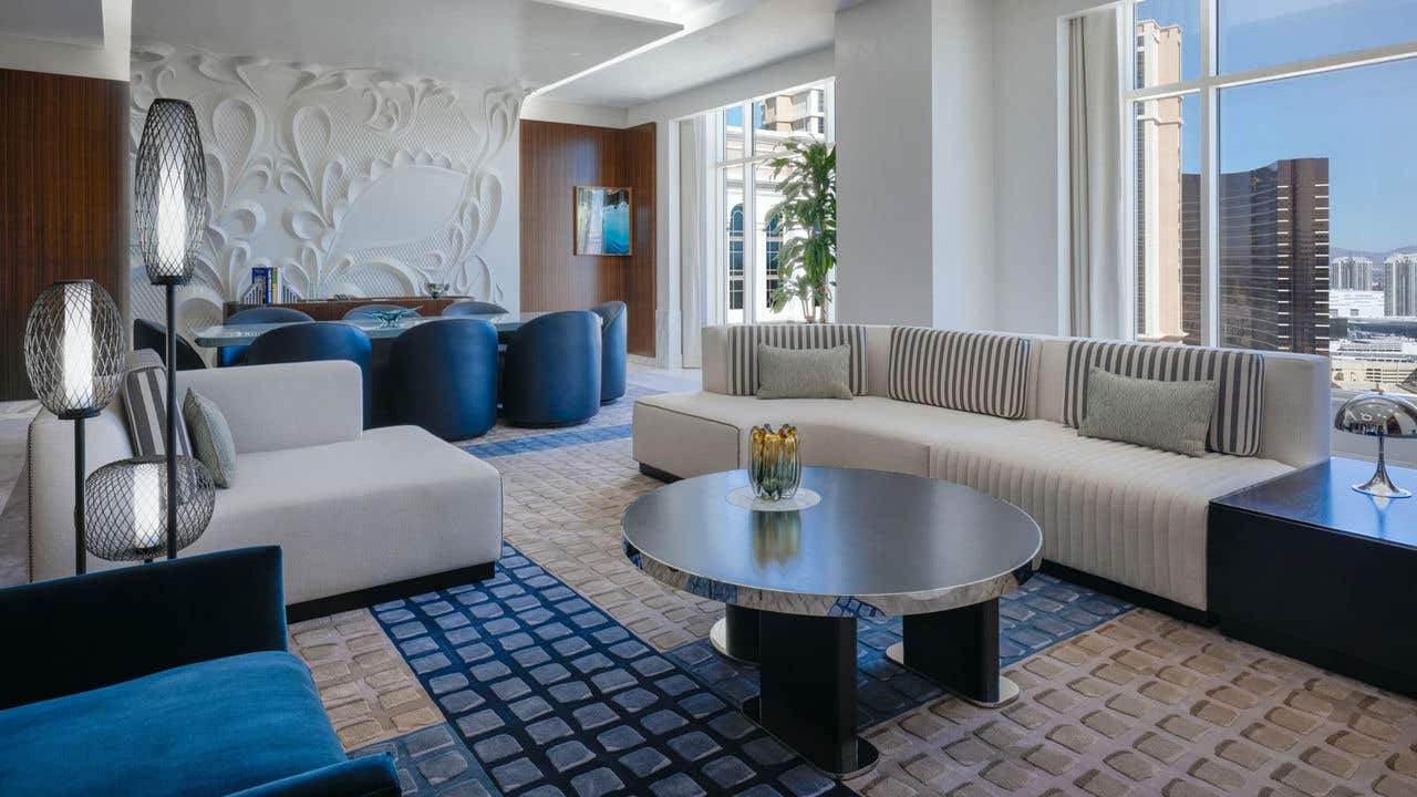 A white and blue hotel living room with a couch, coffee table, chairs, dining table with eight chairs, and two large windows with a view of the Las Vegas Strip.