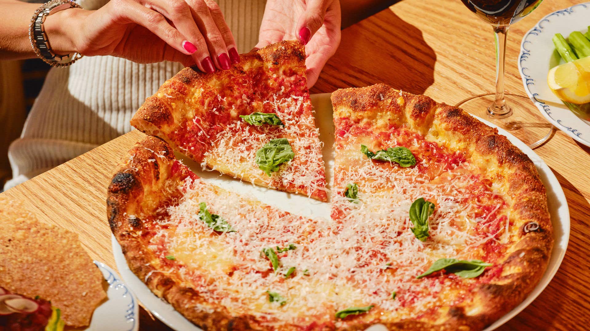 Pizza dish on wood table with hands holding a slice of pizza and a glass of wine nearby.