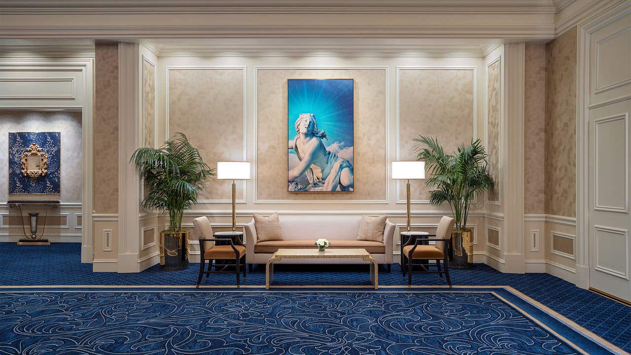 Elegant sitting area with a sofa, chairs, tall lamps, potted plants, and framed artwork, set against ornate walls and blue patterned carpet.