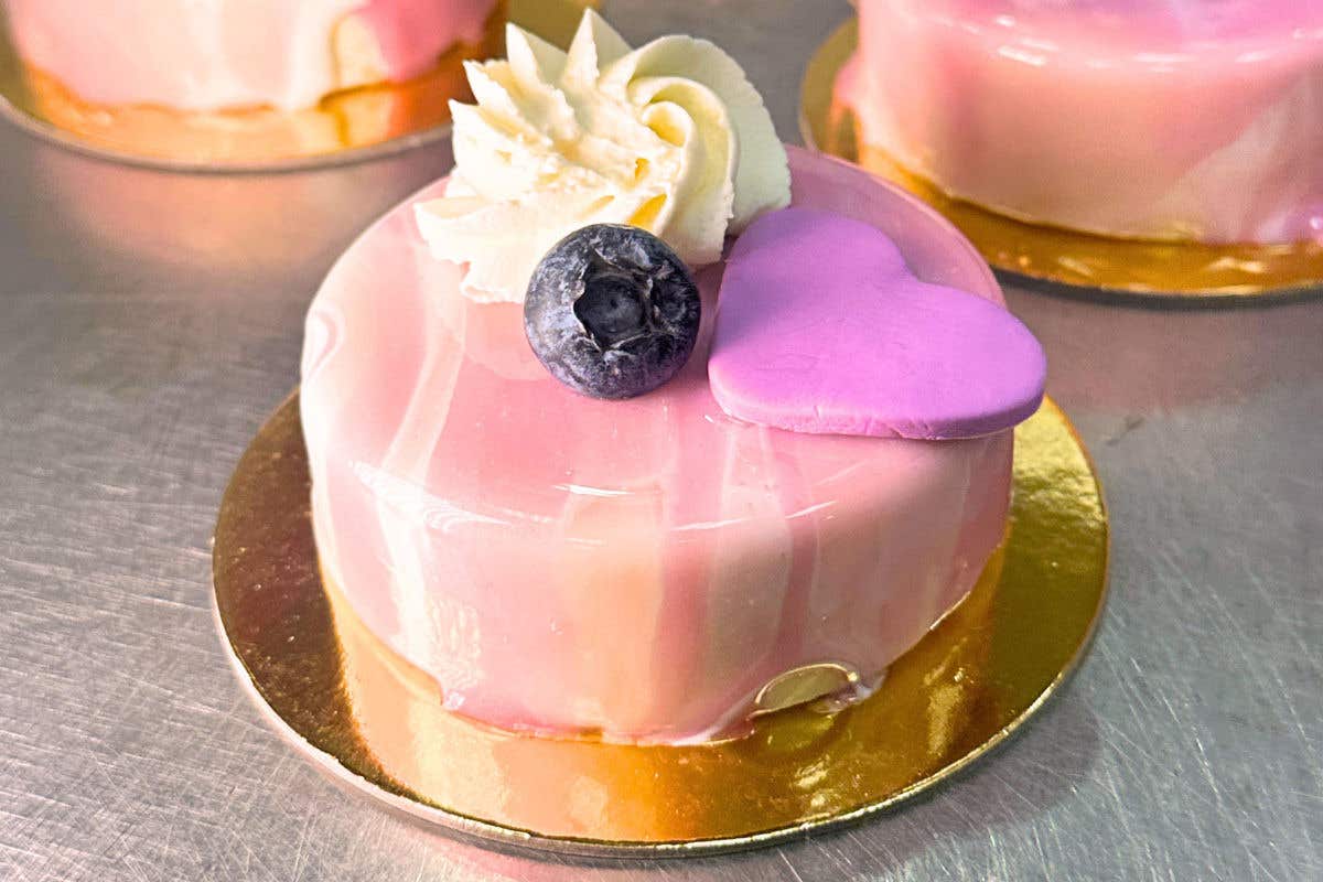 A close-up of a personal-sized dessert with a pink and white marbled mirror glaze, sitting on a gold cardboard base. It is garnished with a rosette of whipped cream, a single fresh blueberry, and a purple candy heart.