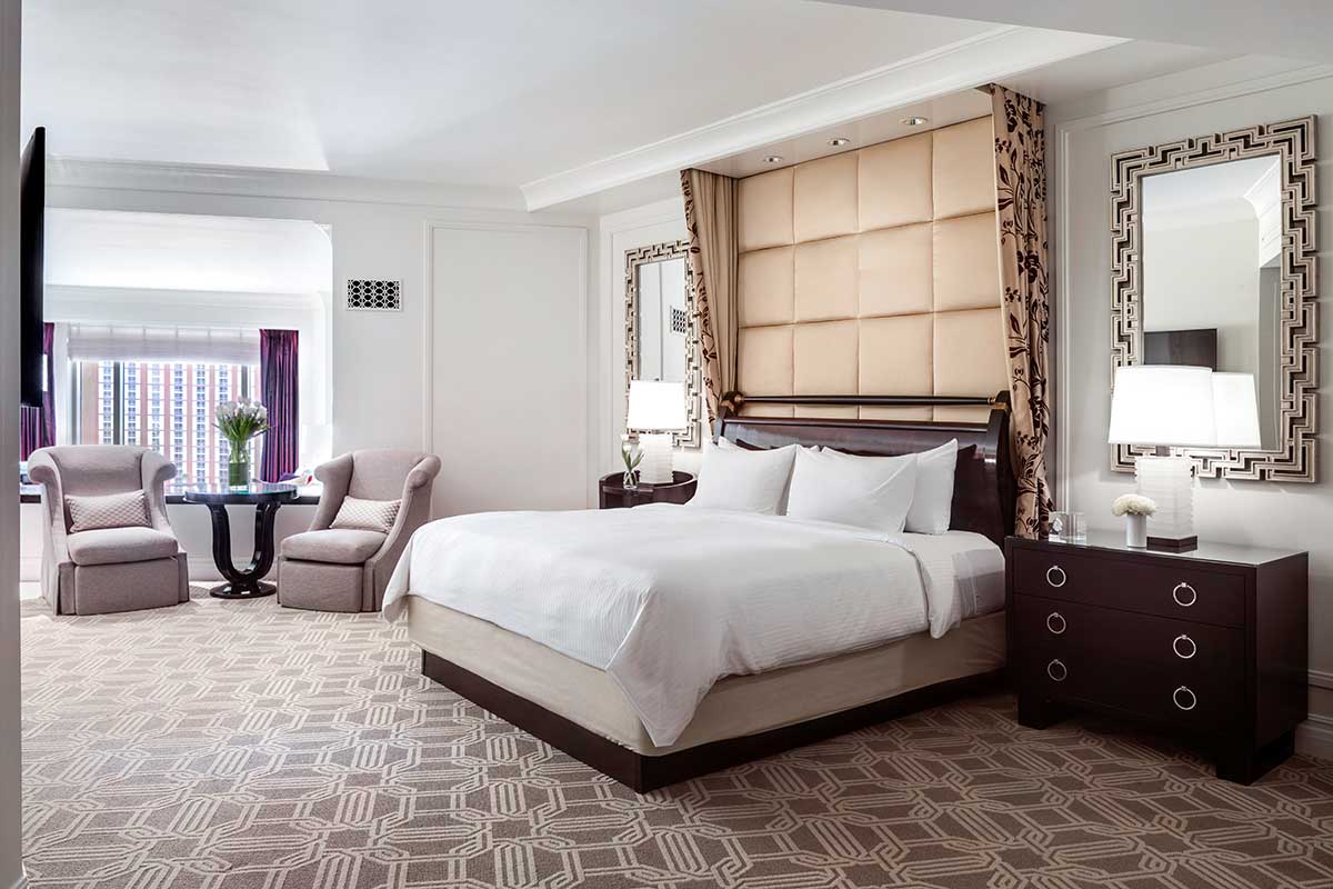 Hotel room with a king bed, plush headboard, two armchairs by a round table, and Strip view from the window. Sophisticated decor with mirrored panels and bedside lamps.