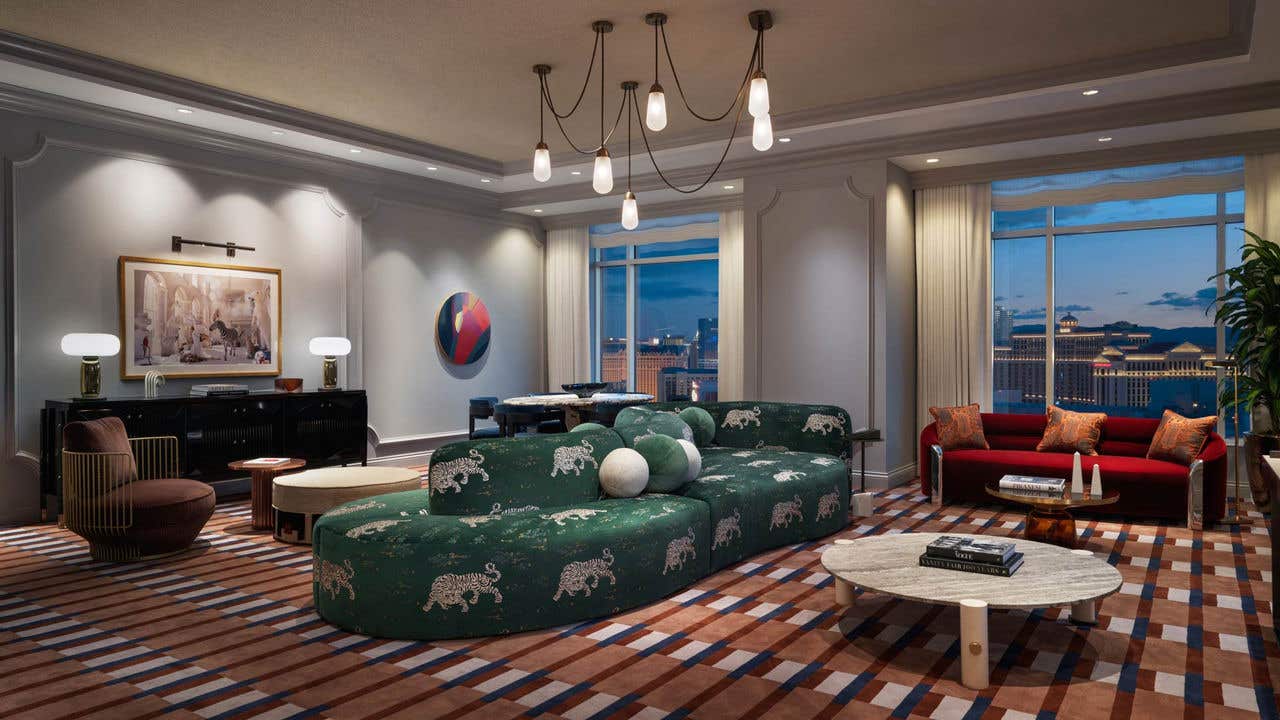 A hotel living room at night with a double-sided dark green couch, chandelier, coffee table, love seat, dining table, armchair, and two large windows with a view of the Las Vegas Strip.