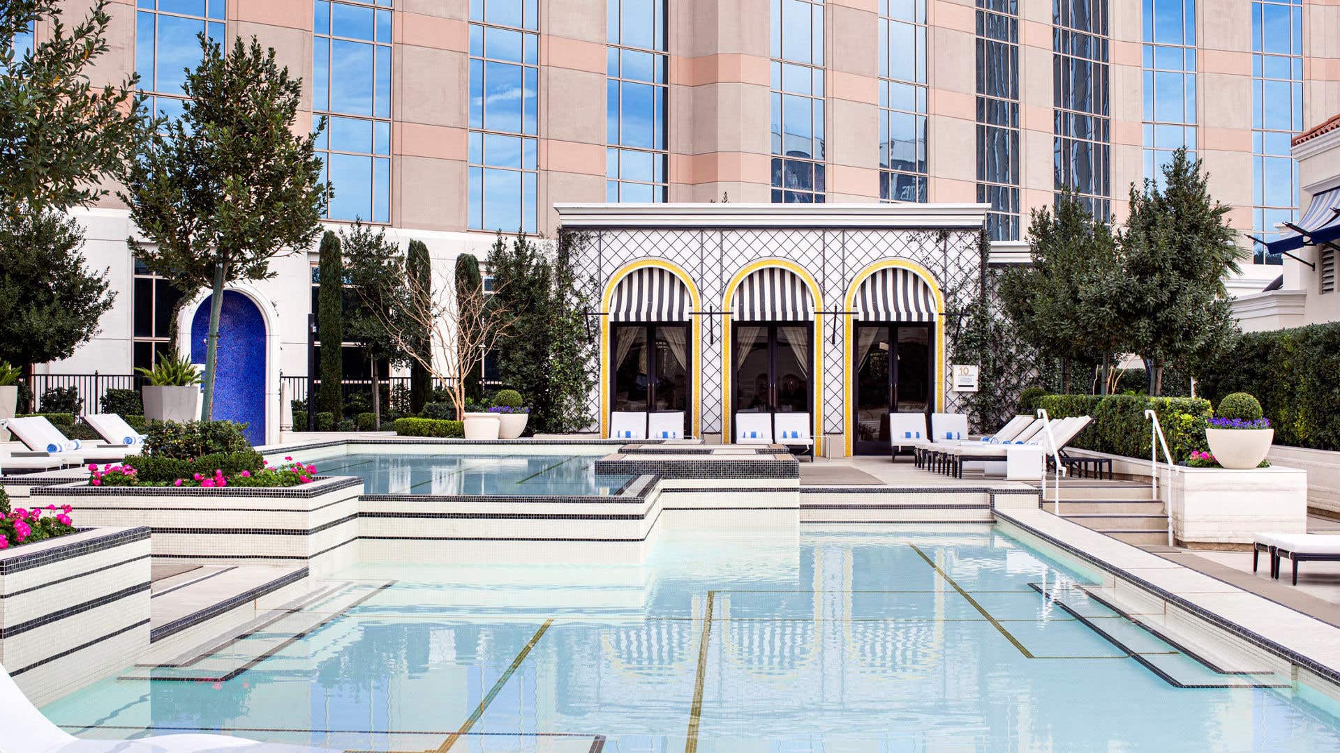 Tiered resort pool with steps leading into the water, bordered by landscaped trees and cabanas with striped awnings in front of a hotel building.