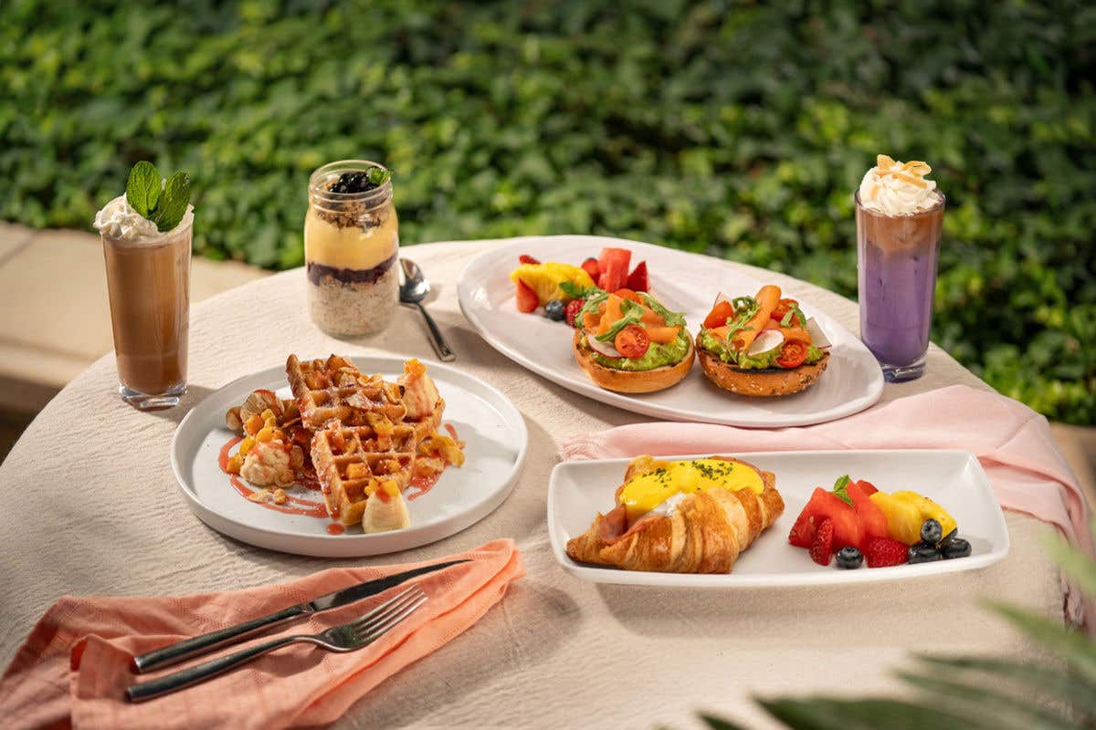 A table set outdoors with assorted breakfast dishes, including waffles, a croissant eggs Benedict, smoked salmon bagels, fresh fruit, a parfait, and two iced drinks.