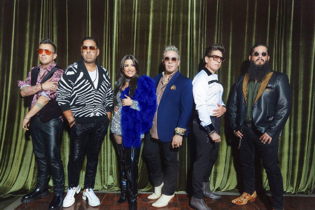 A group of six people standing in front of a green curtain, wearing stylish and colorful outfits.