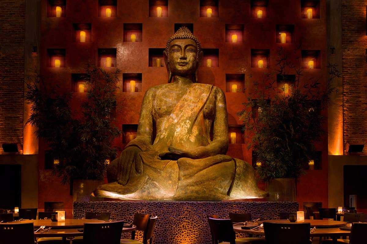 Large seated Buddha statue illuminated, placed against a patterned wall with candles and surrounded by dining tables.