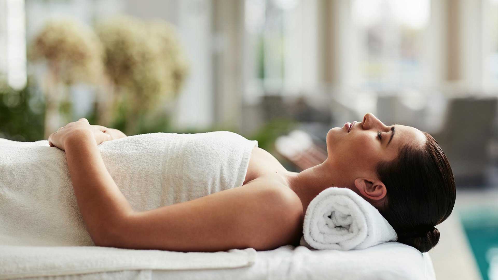 A person lies on a spa table covered in white towels, with a rolled towel supporting the head in a calm spa setting.