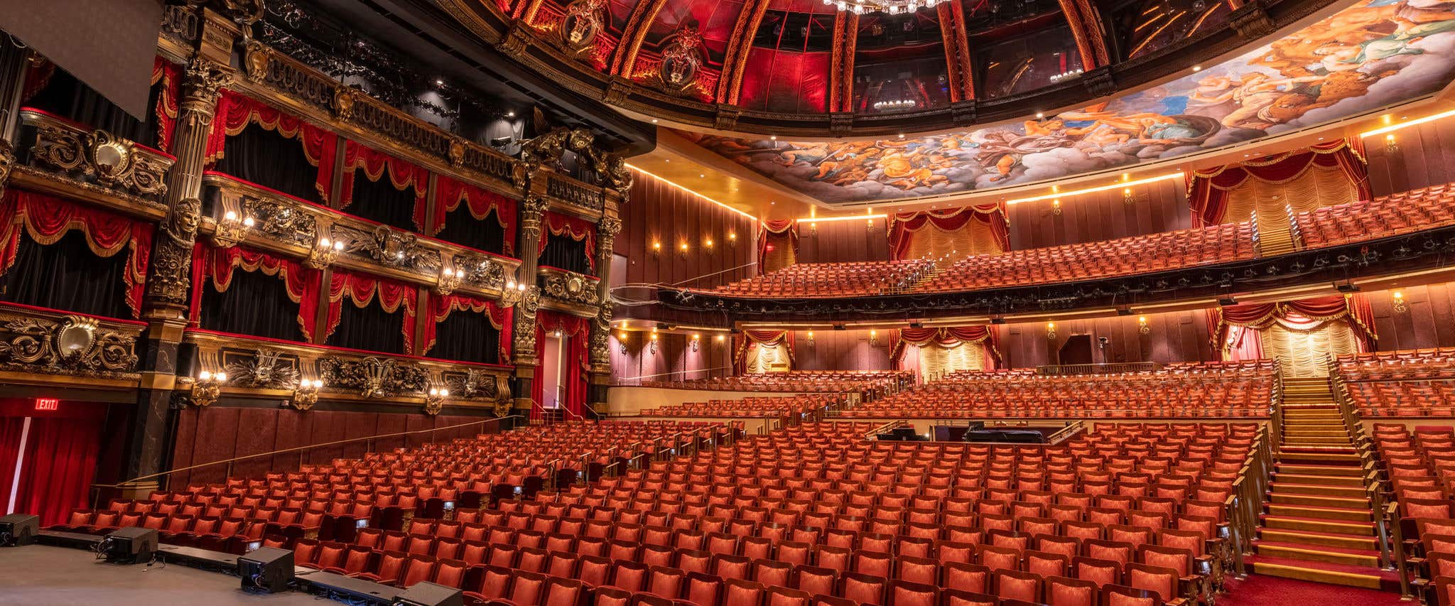 “Wide view of an ornate, Venetian Theatre interior with rows of red upholstered seats facing a stage, gilded balconies on the left, and a painted ceiling mural above, all lit by warm golden lighting.