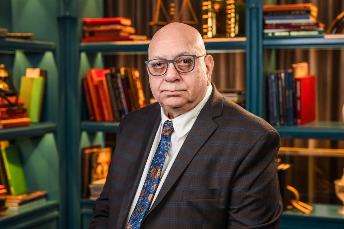 A professional headshot of a man wearing glasses and a dark plaid suit jacket over a white shirt and blue floral tie. He is standing in front of a blue bookshelf filled with books and vintage decor.