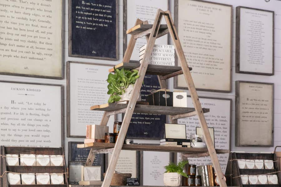Decorative wooden ladder shelf holding plants, candles, and books, with framed text pages on the wall behind.