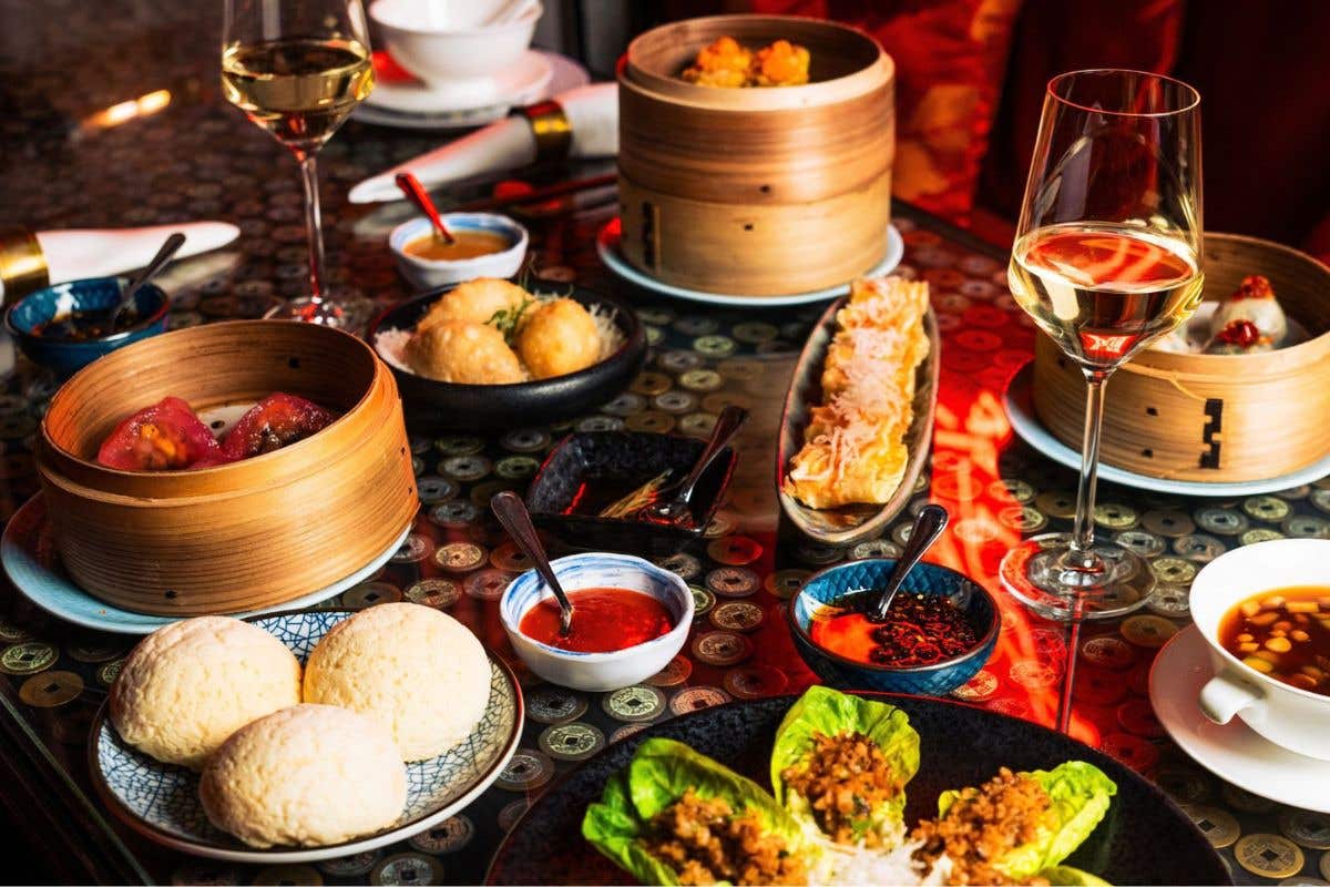 Table spread with dim sum, soups, buns, lettuce wraps, and glasses of wine.