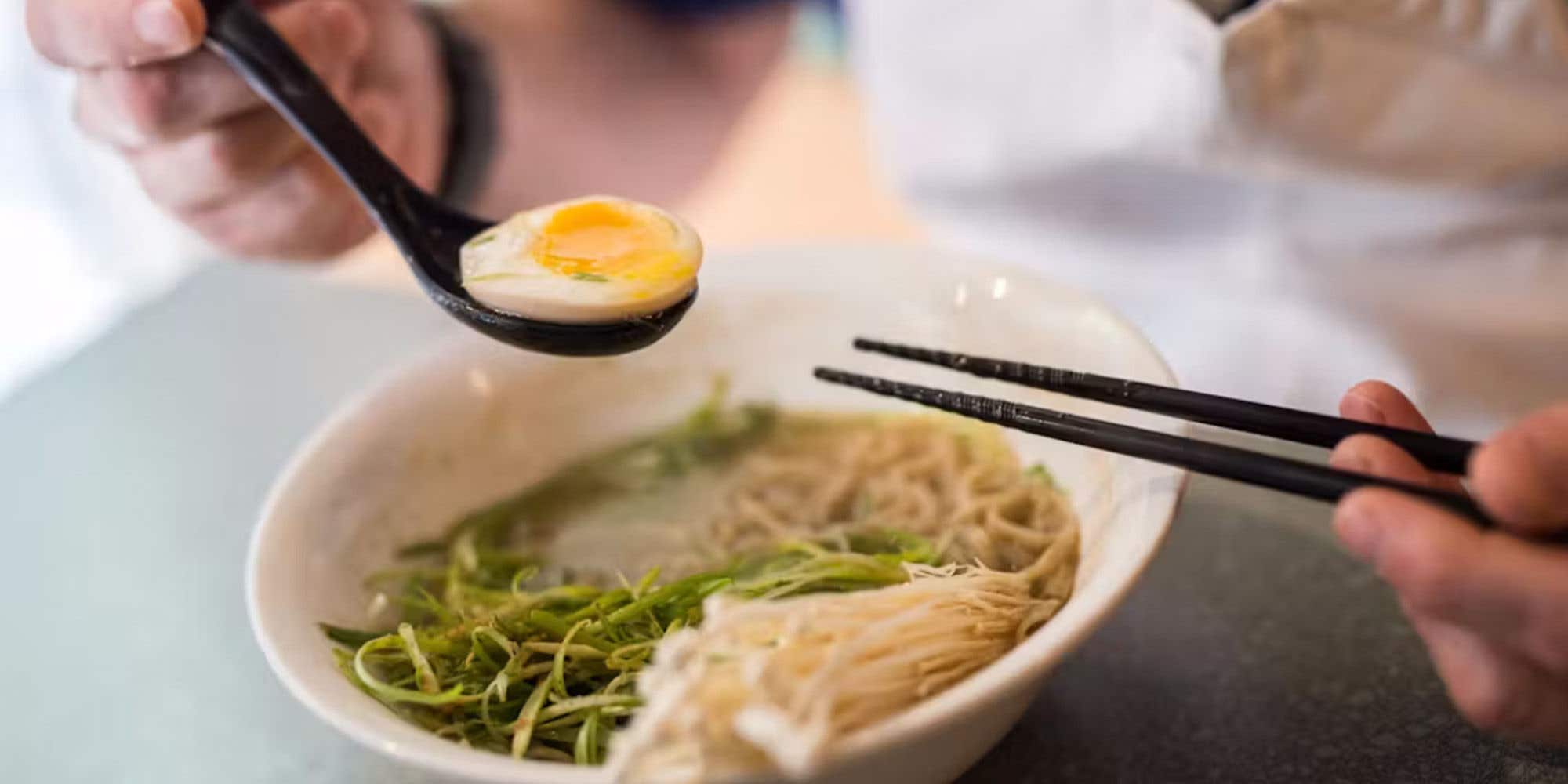 Ramen soup in a bowl with someone holding a hardboiled egg in a spoon and chopsticks over the bowl.