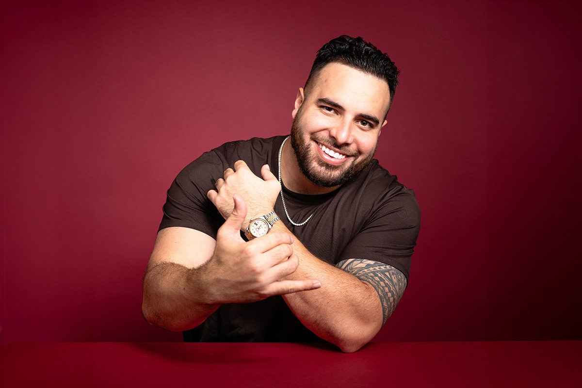 Comedian Tumua smiling, resting on one arm, set against a red background.