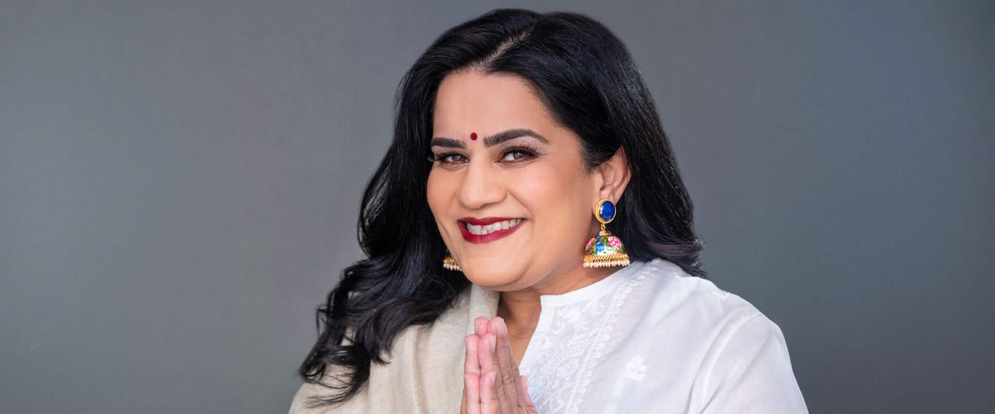 Zarna Garg in a white and beige outfit with her hands together smiling at the camera. 