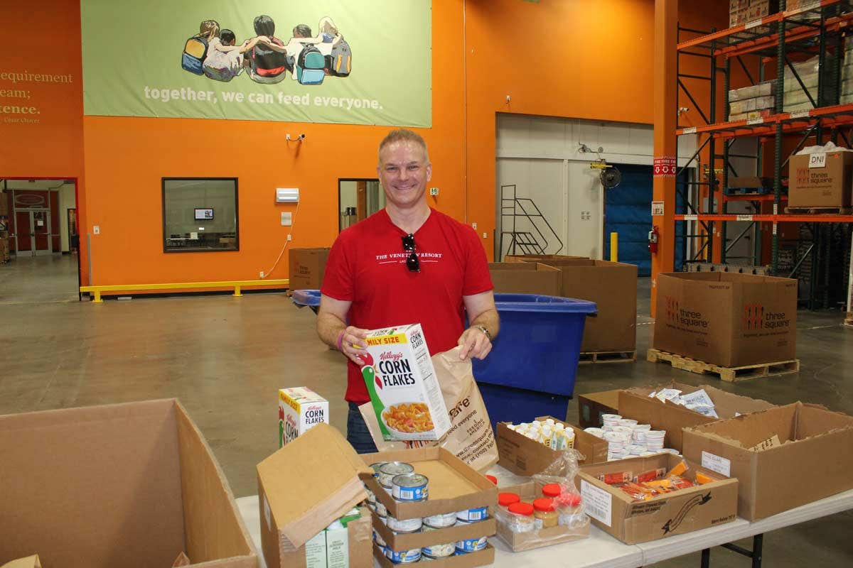 A person in a warehouse smiles while packing food items into boxes, with colorful shelves and a green wall mural in the background.