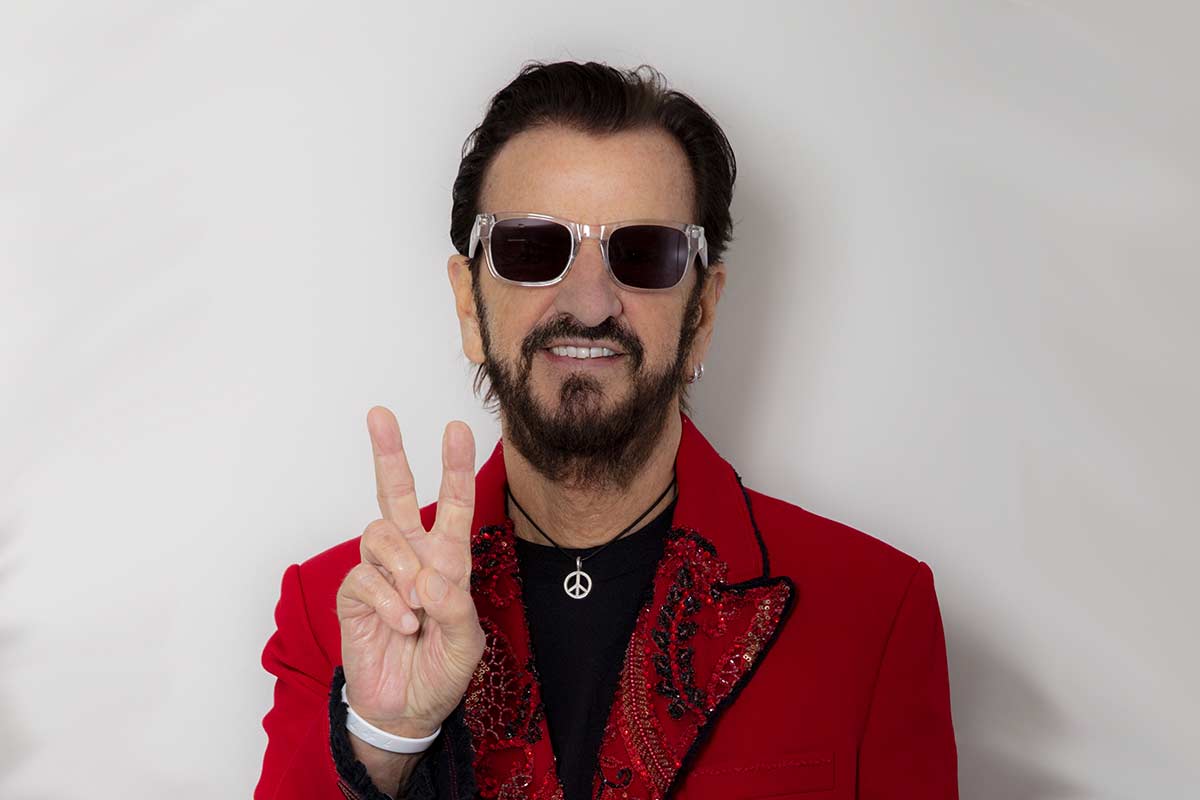 Man in a red jacket and sunglasses making a peace sign with his hand against a white background.