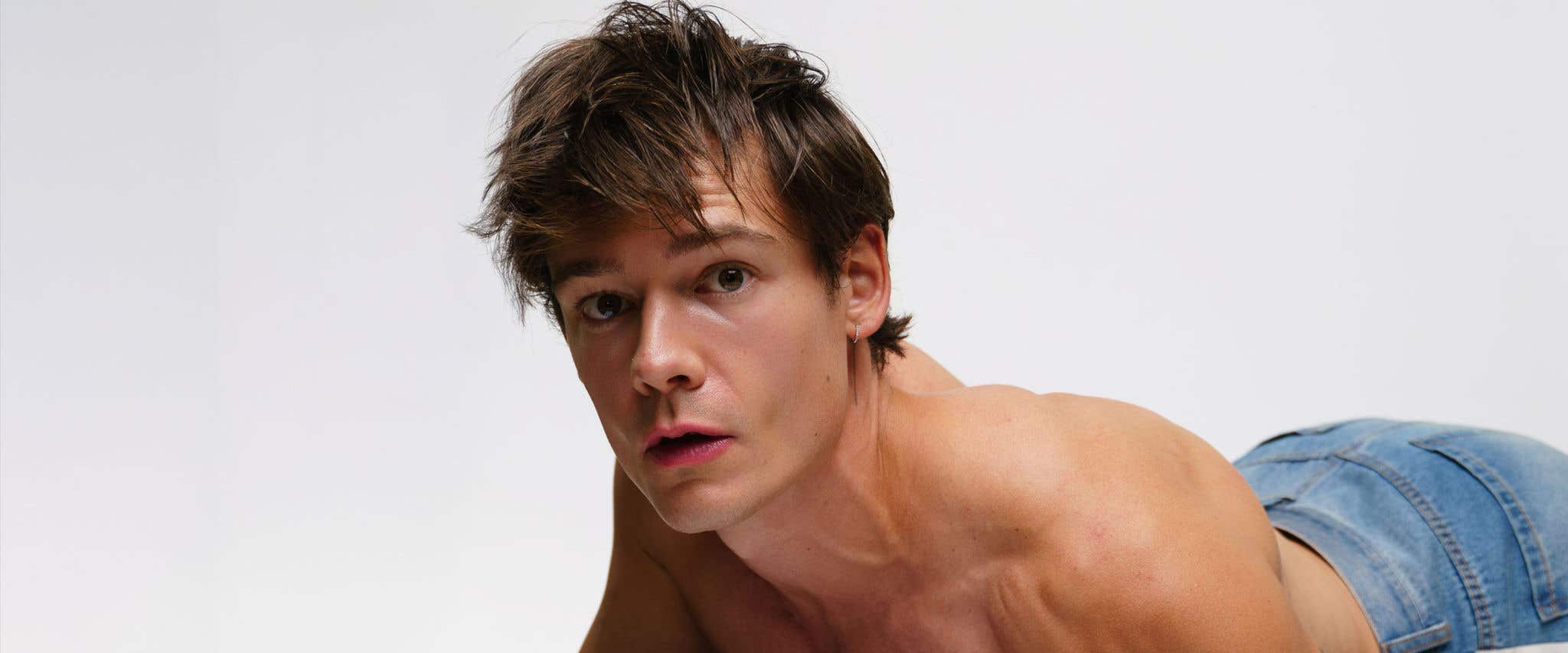 Mario Adrion in jeans propped up off the ground with his forearm with a denim shirt in front of him staring at the camera in front of a white background.