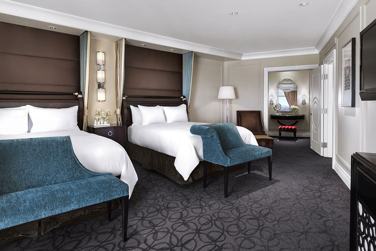 A hotel room with two Queen beds with brown fabric headboards and benches at the end of the bed, a lamp and chair in the corner, and an open doorway to a makeup vanity area on the right. 