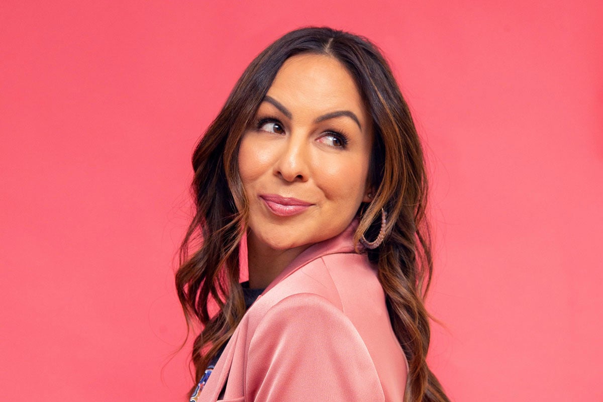 Anjelah Johnson-Reyes in a pink jacket, looking over her shoulder, with a pink backdrop