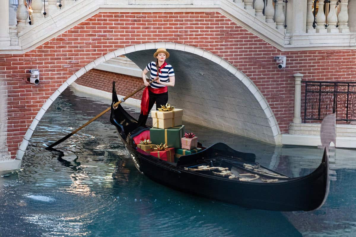 Gondola carrying wrapped gift boxes through a canal beneath a brick bridge at The Venetian.