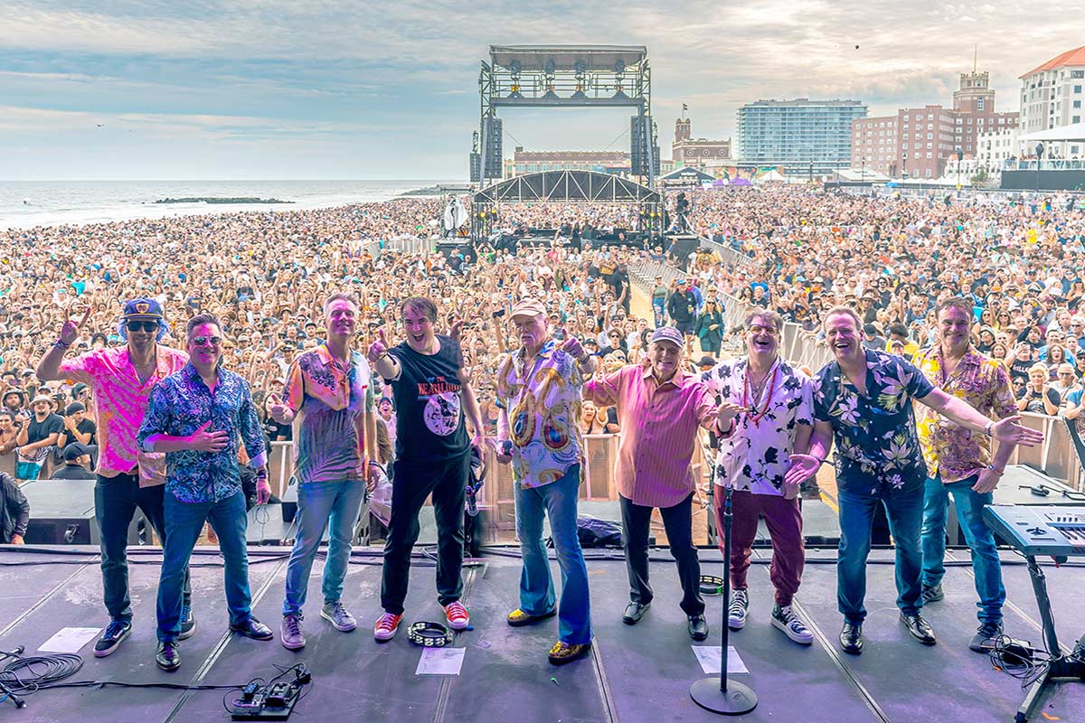 The Beach Boys, standing on stage facing the camera, with a large crowd of concert goers behind them.
