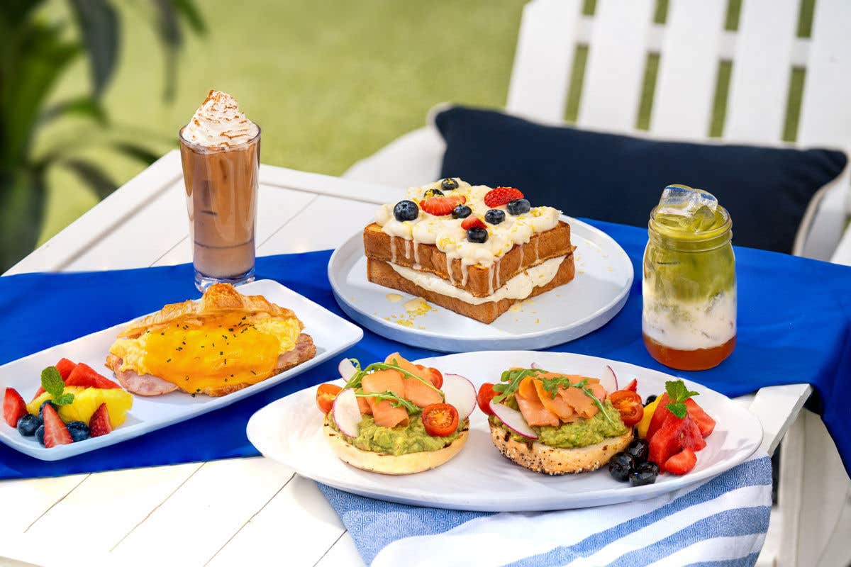 A white outdoor table displays breakfast dishes, including croissant eggs Benedict, avocado and salmon bagels with fruit, a stack of waffles topped with cream and berries, an iced chocolate drink, and a layered iced matcha drink.