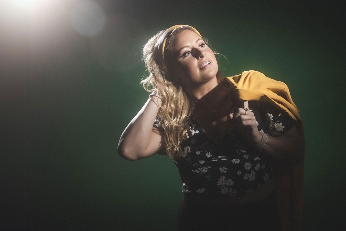Becky Robinson in a black floral top with a yellow headband and jacket against a dark green background.