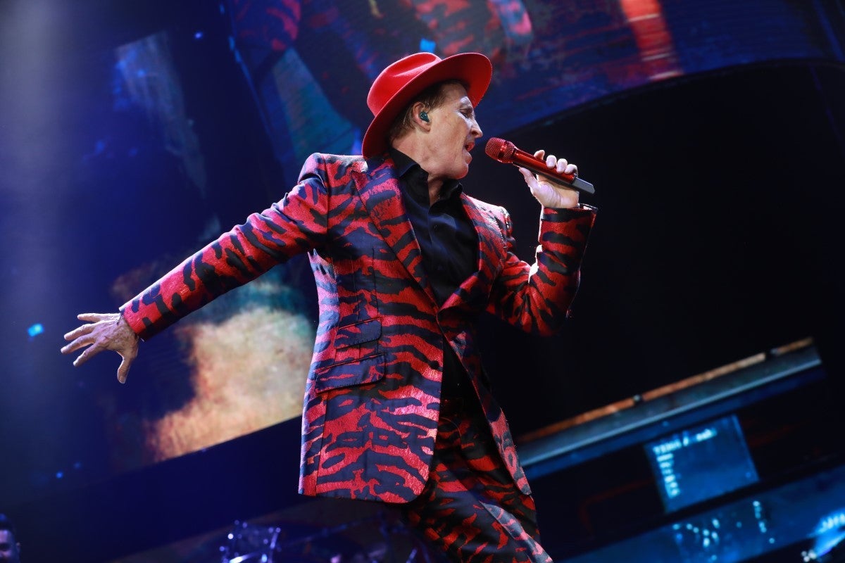 Person in a red and black patterned suit and hat singing on stage with a microphone.