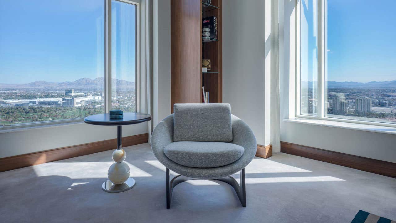 An armchair, side table, and bookshelf sit in the corner of a room with two large windows on both walls with a high-rise view of the Las Vegas valley.