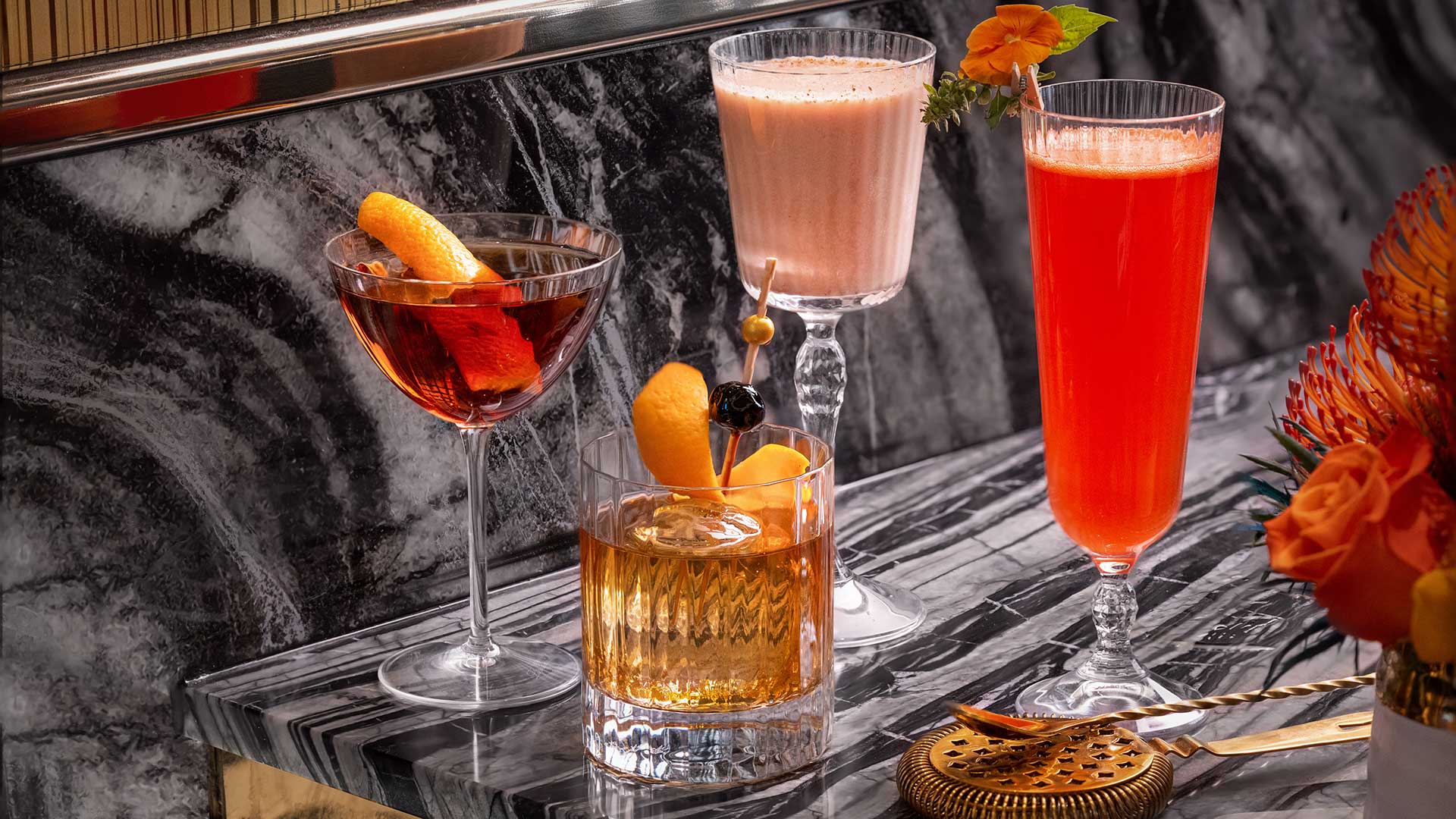 three cocktail glasses on a marble bar with flowers and orange garnish