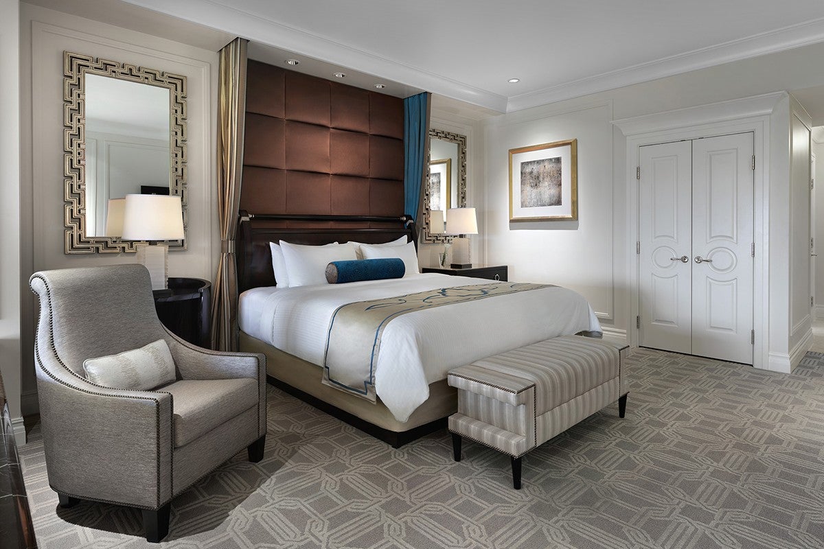 Modern hotel room with a large bed, upholstered headboard, armchair, bench, and mirrored nightstands. Neutral colors.