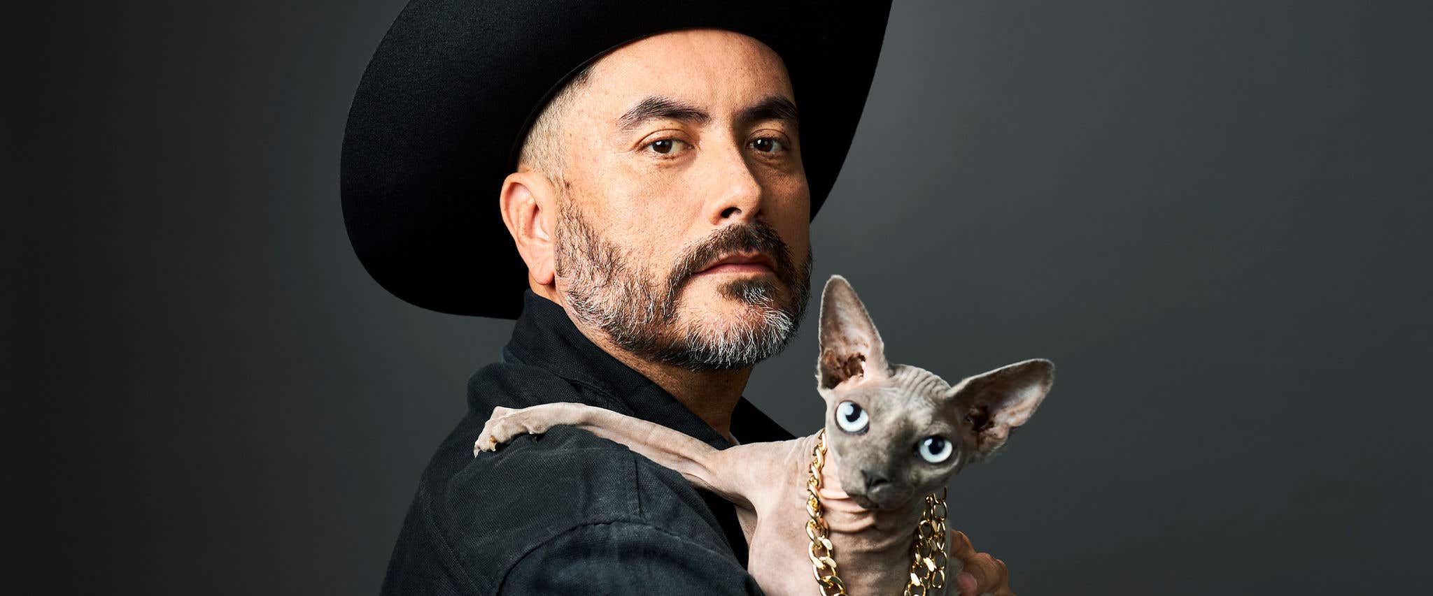A man in a black cowboy hat holds a Sphynx cat wearing a gold chain necklace.