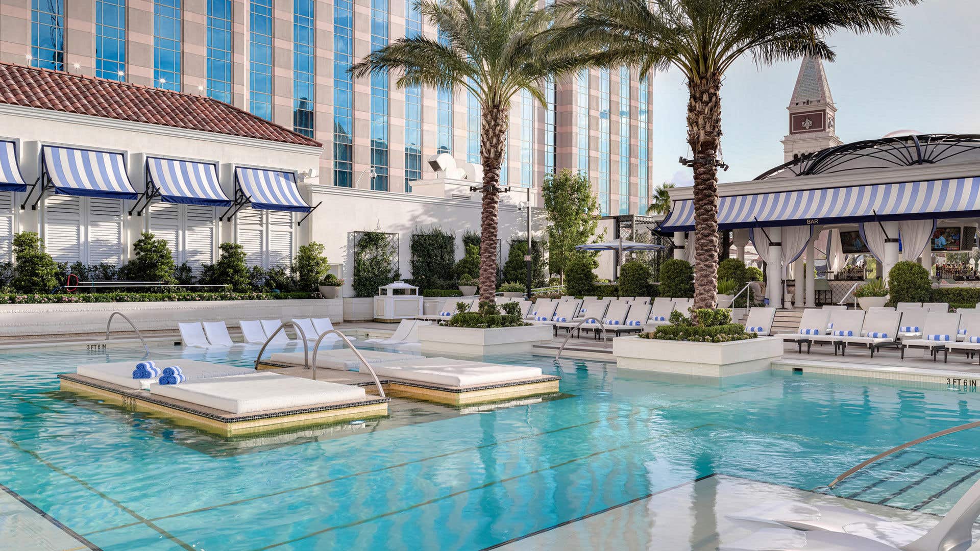 Resort pool with submerged white loungers and railings, bordered by palm trees and striped cabanas, with a tall glass hotel tower in the background.