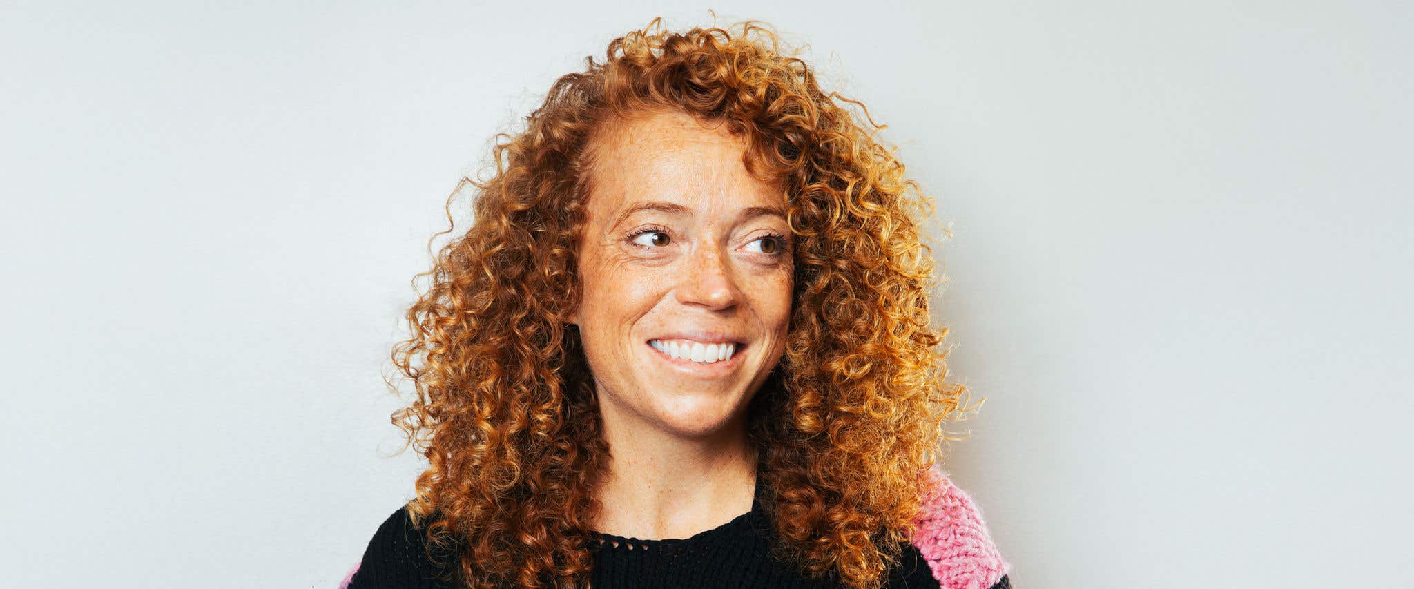 Michelle Wolf in a black and pink knit sweater looking off into the distance in front of a gray background.