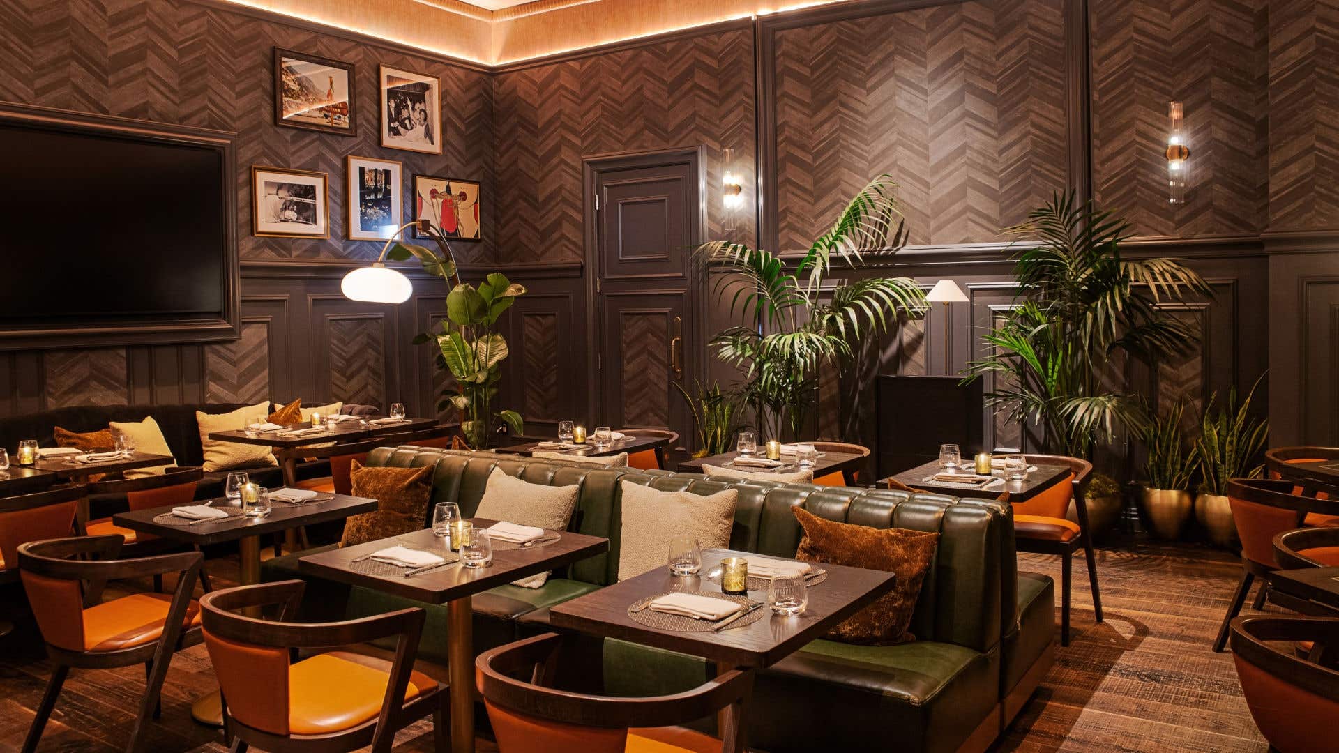 Upscale speakeasy with leather banquettes, warm lighting, wood-paneled walls, framed art, and lush indoor plants.