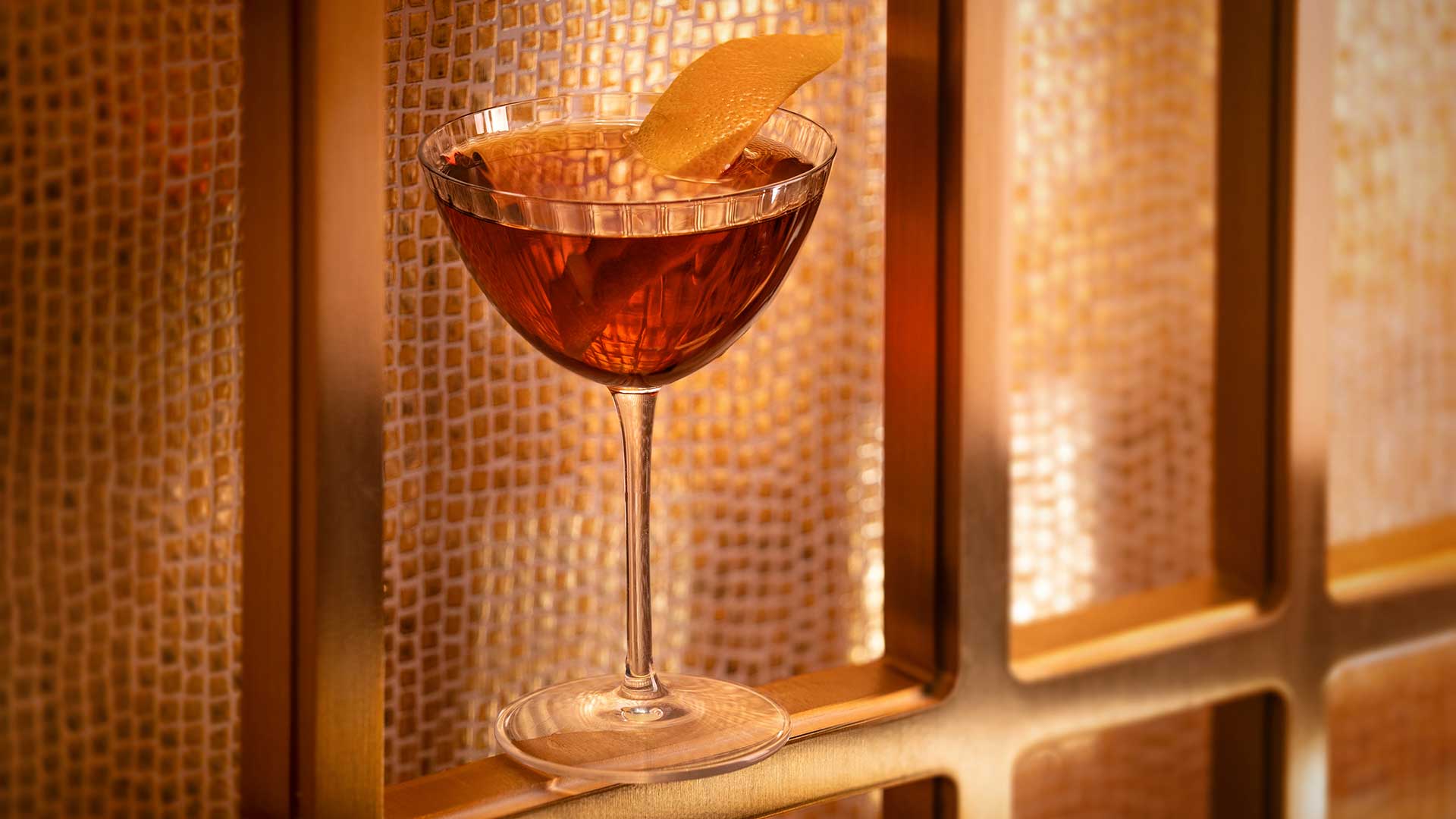 A cocktail glass with a brown drink and lemon peel garnish sits on a ledge in front of a gold mosaic tile wall.
