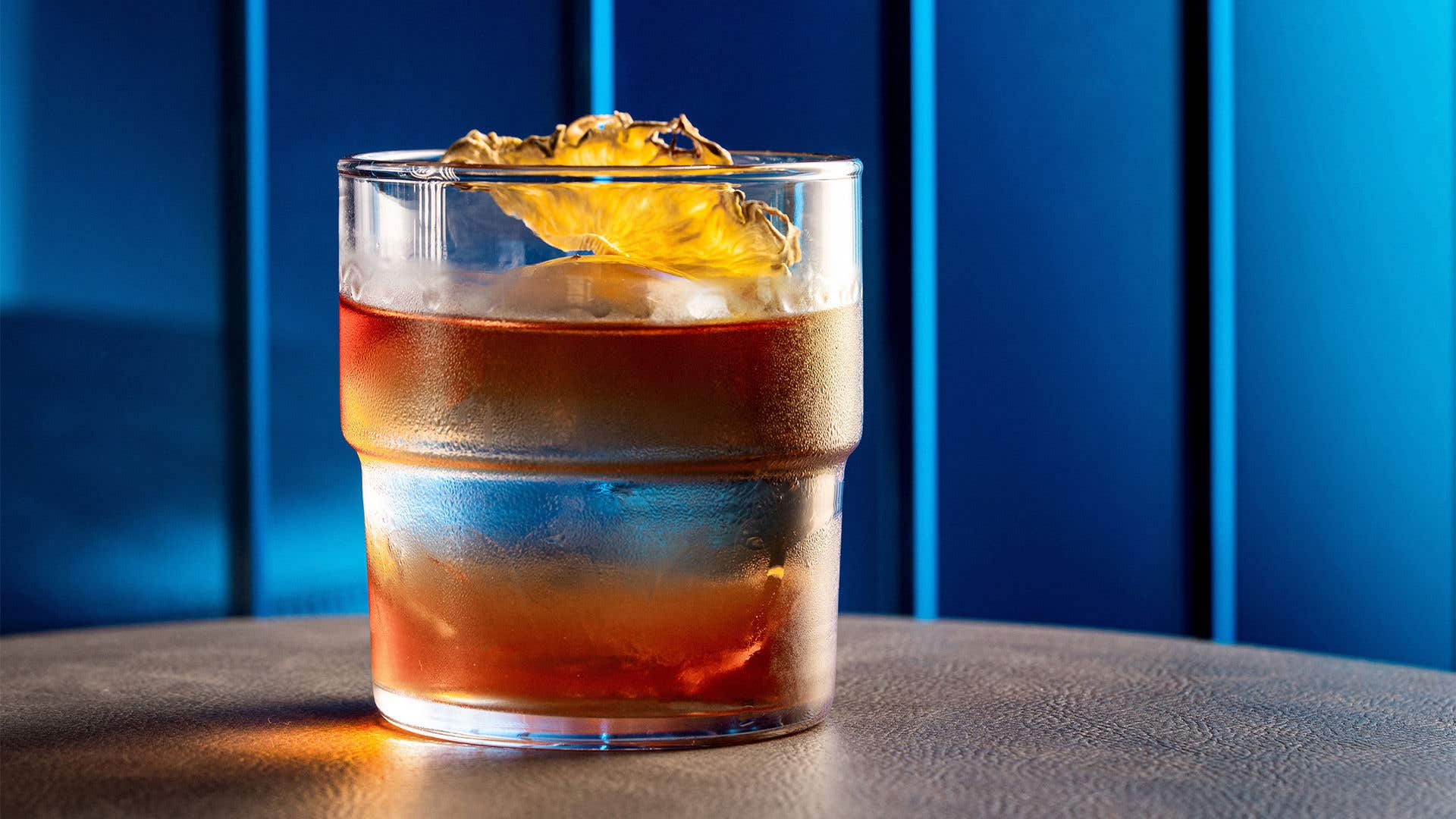Cocktail with layered orange and clear liquor in a small glass garnished with dried orange slice. 