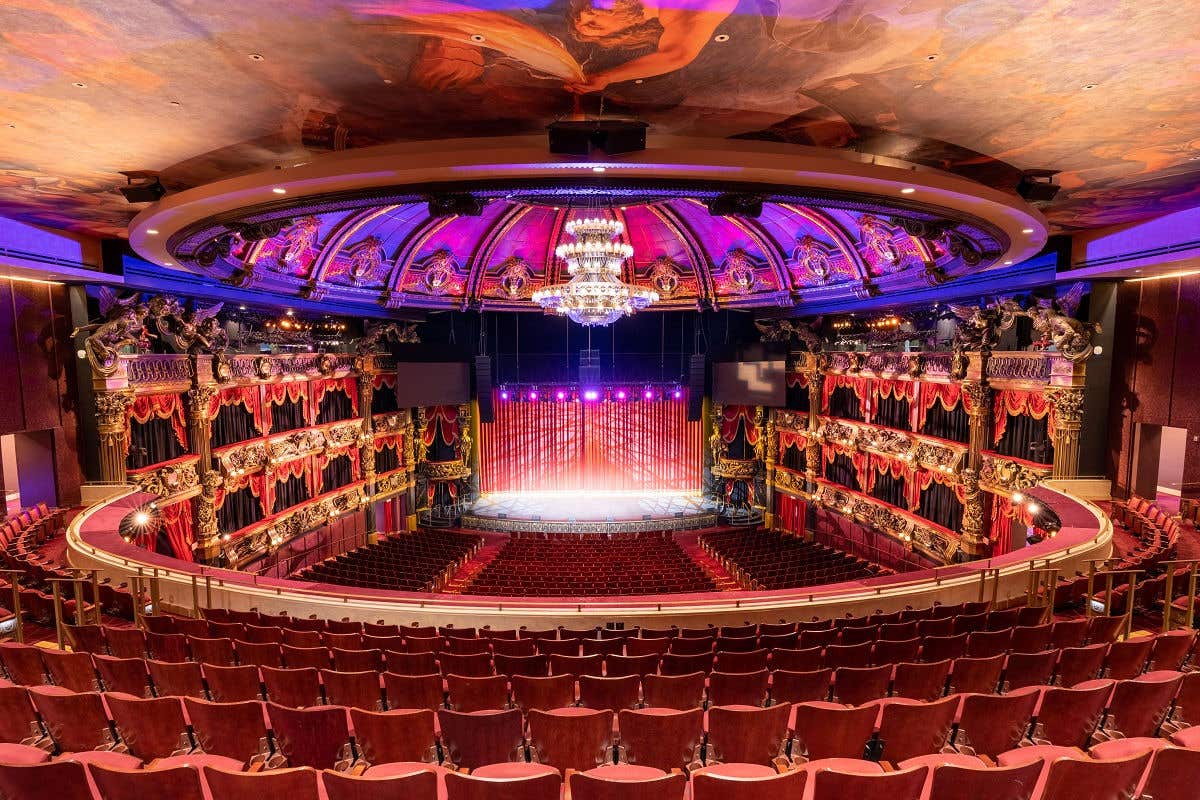 Ornate theater interior with a grand chandelier, red velvet seating, and an illuminated stage, surrounded by detailed balconies and murals on the ceiling