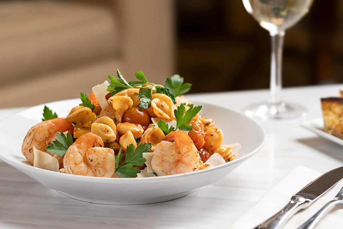 A bowl of shrimp pasta with parsley and a glass of white wine on a table.