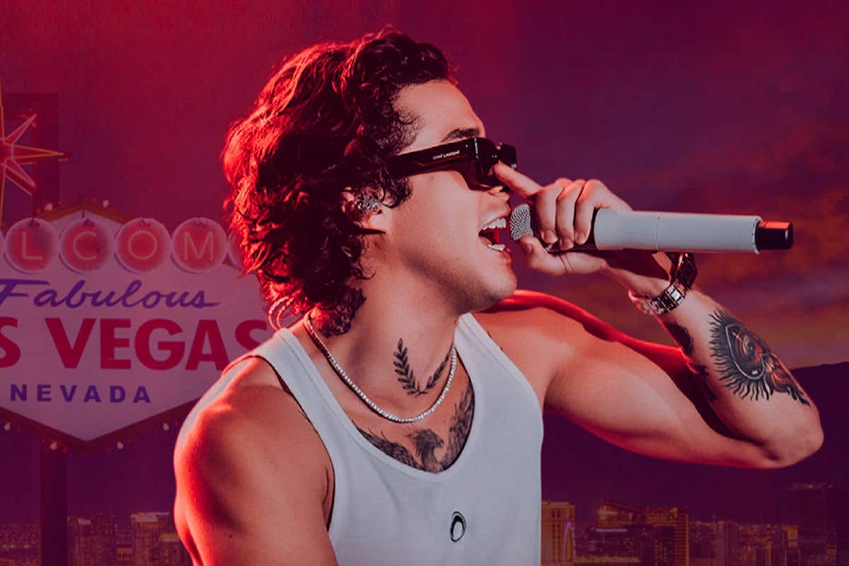 Mario Bautista in a white tank and black bottoms holding a microphone and pushing up dark sunglasses standing in front of Welcome to Fabulous Las Vegas sign.     