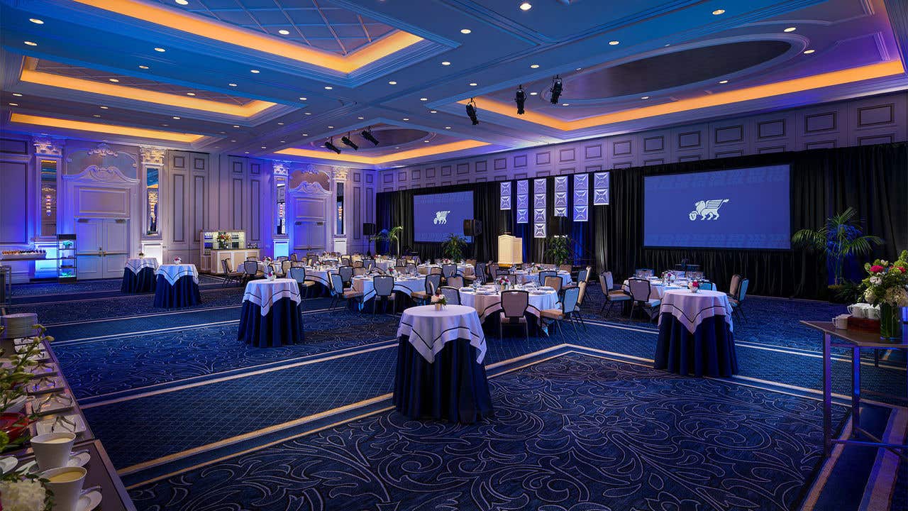 Elegant ballroom with round tables in navy and white linens, blue accent lighting, two projection screens, and a staged podium.