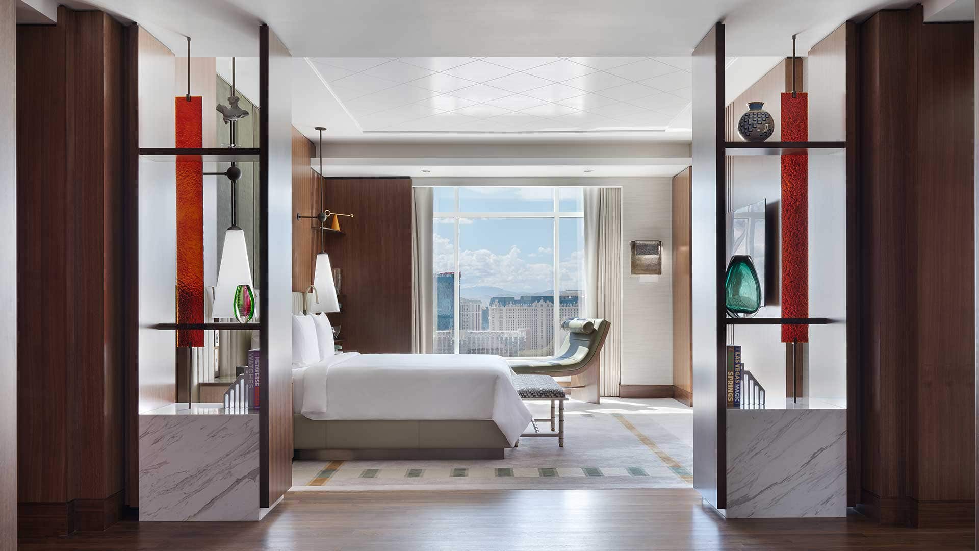 A bedroom with a king bed, chaise lounge next to the window, decorative bookshelves, and a view of the Las Vegas Strip.