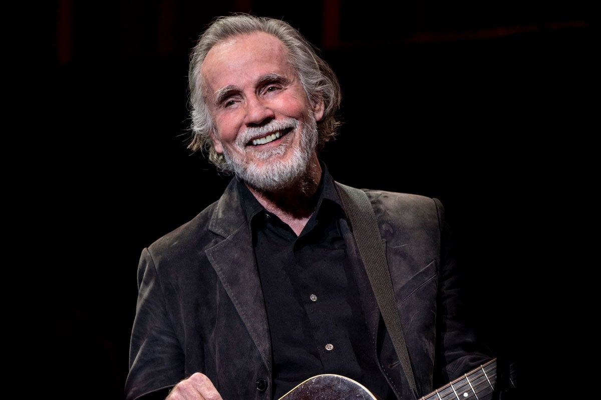 Jackson Browne, in dark attire, smiling and holding a guitar against a black backdrop