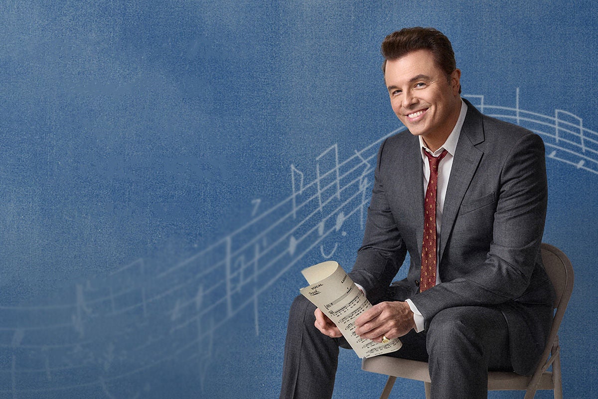 Actor and singer Seth MacFarlane sitting on a folding chair, set against a blue background with music notes.