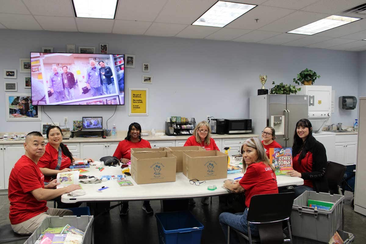 Seven people in red shirts working together in a room with boxes and books. A TV is on the wall showing a video.