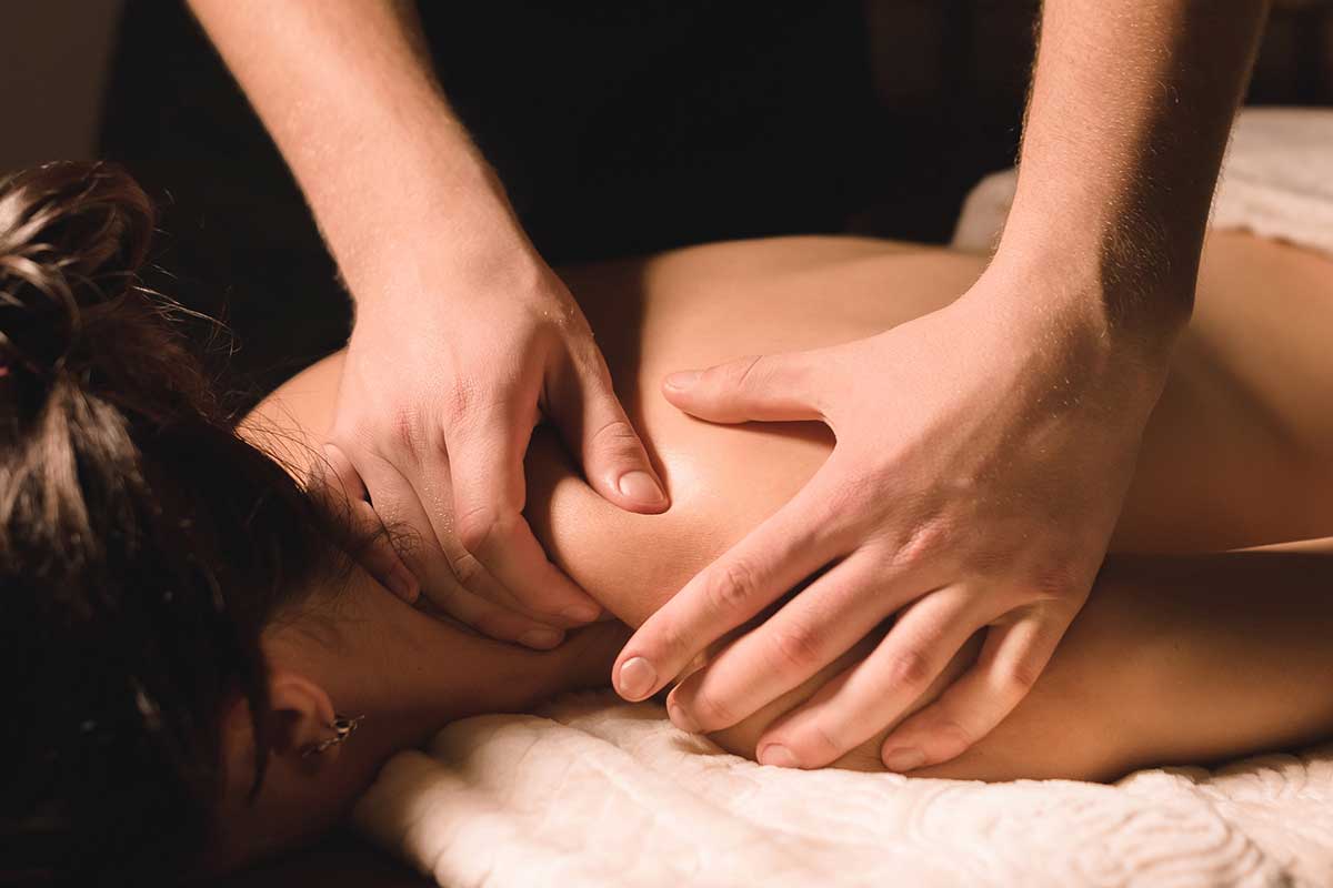 A massage therapist uses both hands to apply pressure to a person’s upper back and shoulder while they lie face down on a padded table.