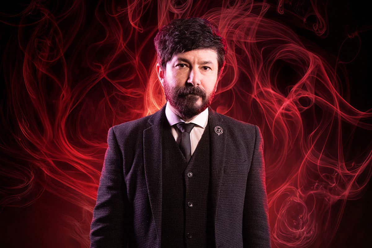 Stuart Macleod in black suit jacket, black vest, black tie and white shirt standing in front of maroon colored smoke on black background