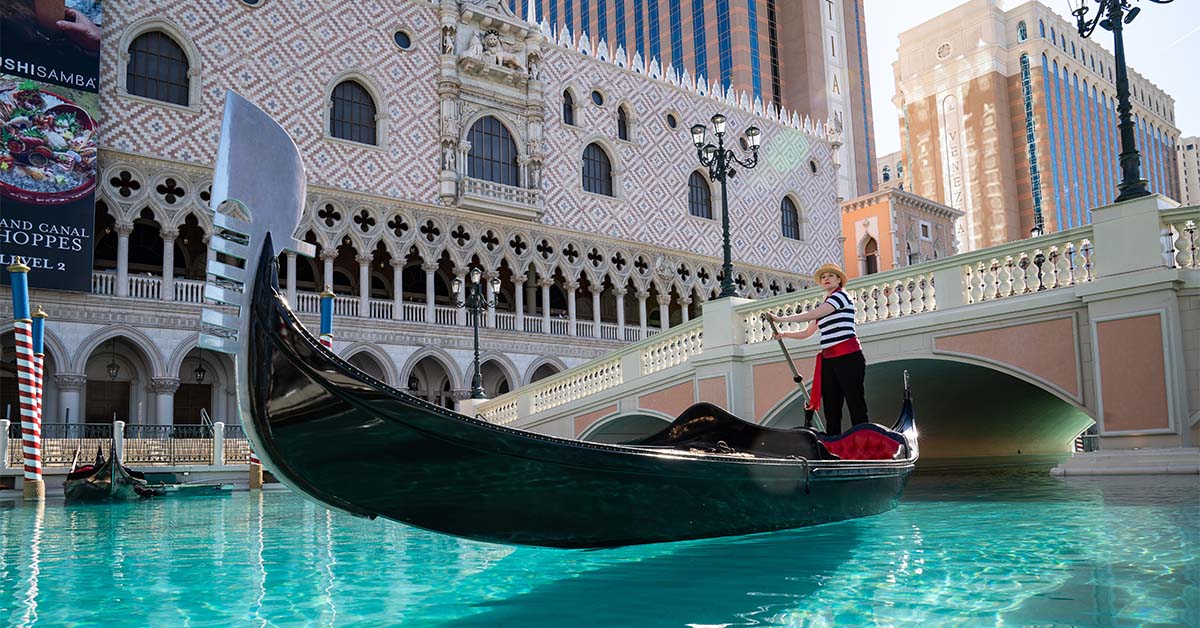 A gondolier steers a gondola across a canal in front of a large building with a bridge.