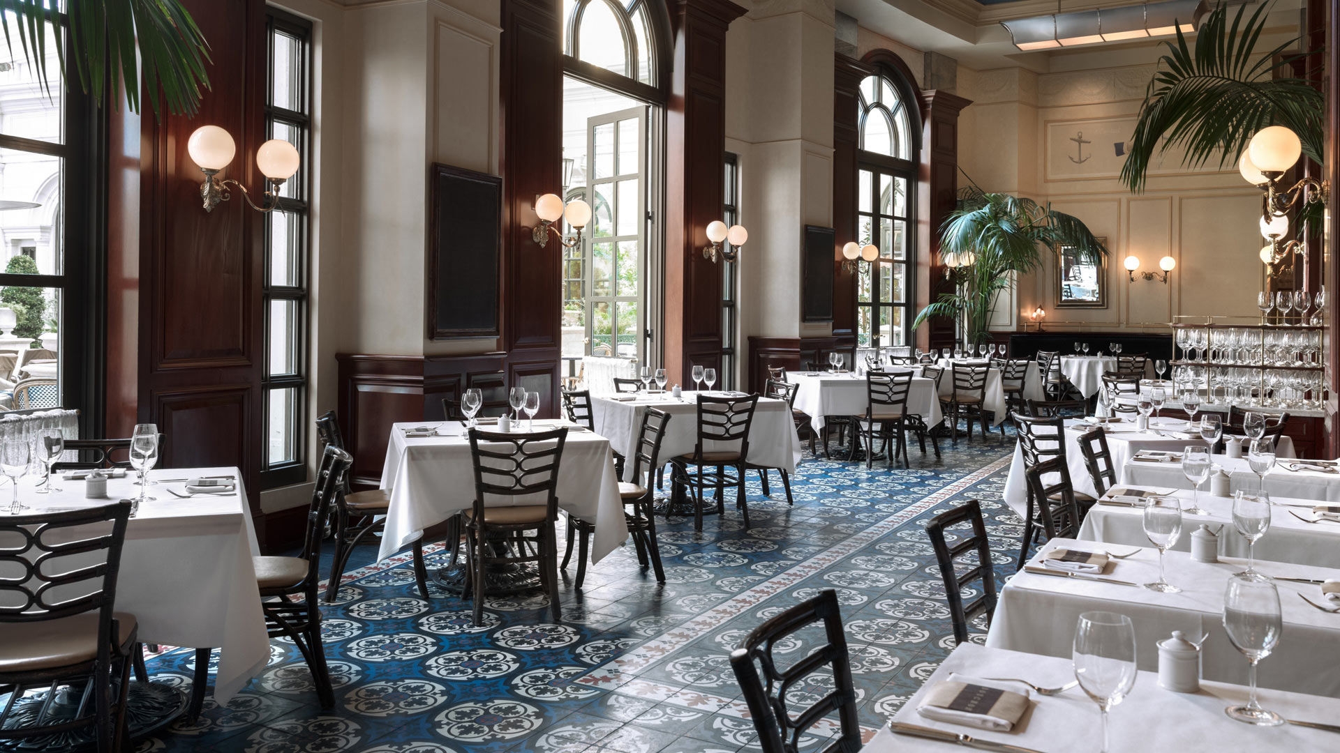 Elegant restaurant dining area with white tablecloths, dark wood paneling, large arched windows, and blue patterned floor tiles.
