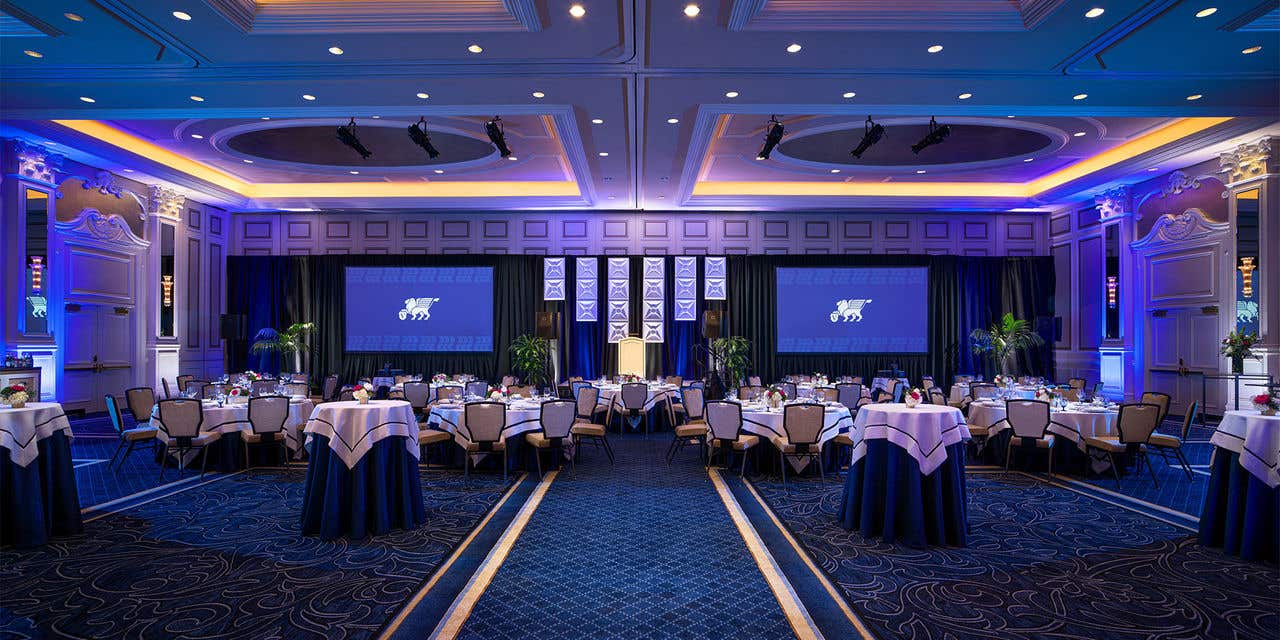 A large, elegant ballroom set for an event, featuring round tables draped in navy and white linens, banquet chairs, and two large projection screens at the front of the room. 